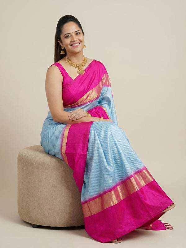 Woman wearing a pink and blue saree sitting on a beige stool against a beige background