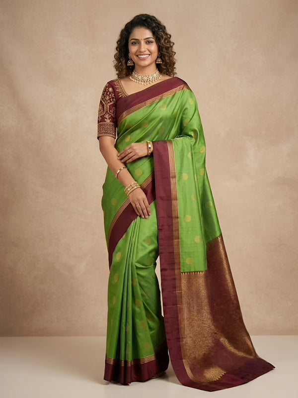 A woman modeling a green silk saree with a deep maroon border and gold zari motifs, paired with a matching embroidered blouse, set against a neutral beige studio backdrop.