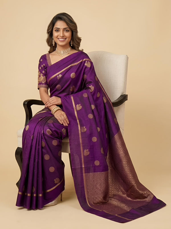 A woman seated on a white armchair wearing a purple silk saree with gold zari motifs and a matching blouse, photographed against a plain beige studio background.