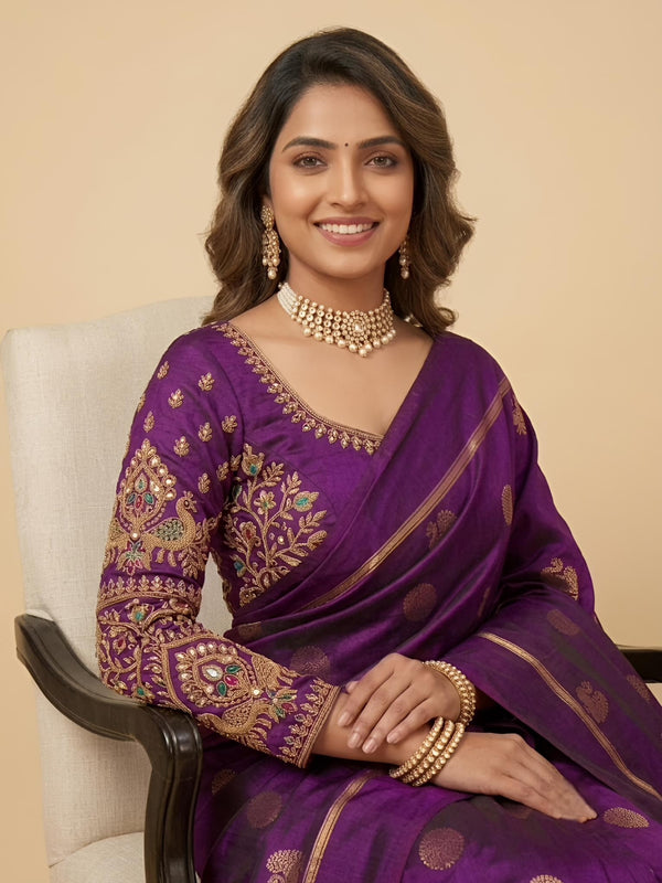 A woman seated in a light upholstered chair wearing a purple saree with ornate gold embroidery and a matching embroidered blouse. She is styled with gold jewelry against a plain beige studio background.